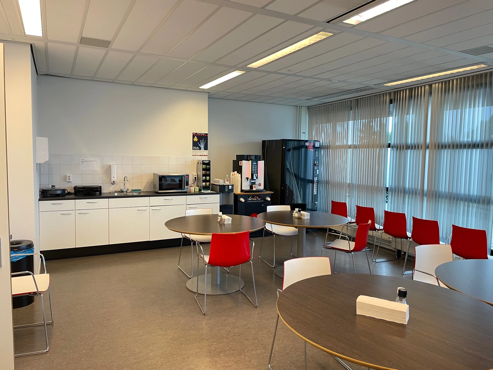 Kantine aan de Karel Doormanweg met ronde tafels, rode stoelen, een kitchenette en koffieautomaat.